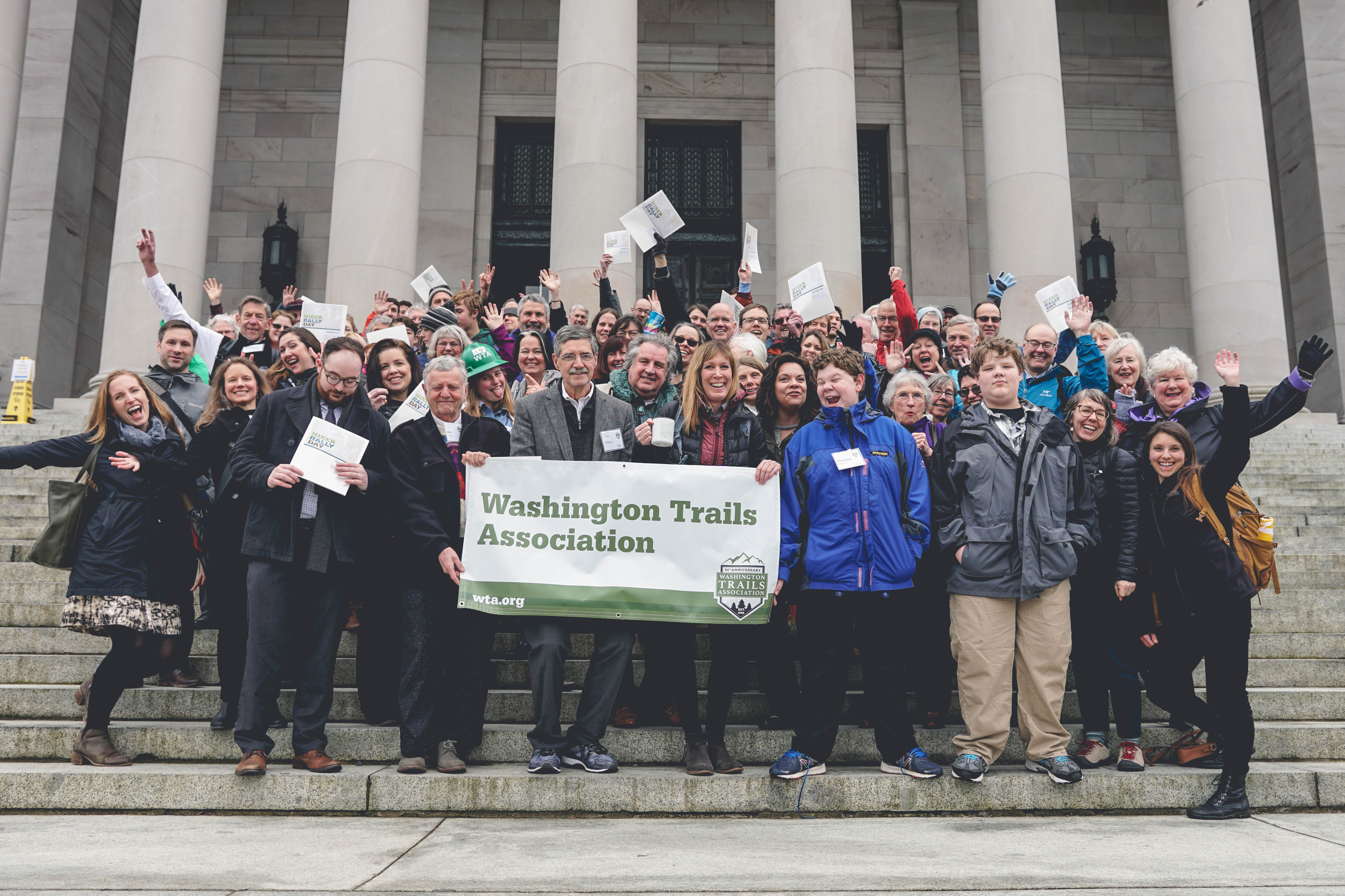 Dozens of hikers, smiling with arms raised, on the capitol steps
