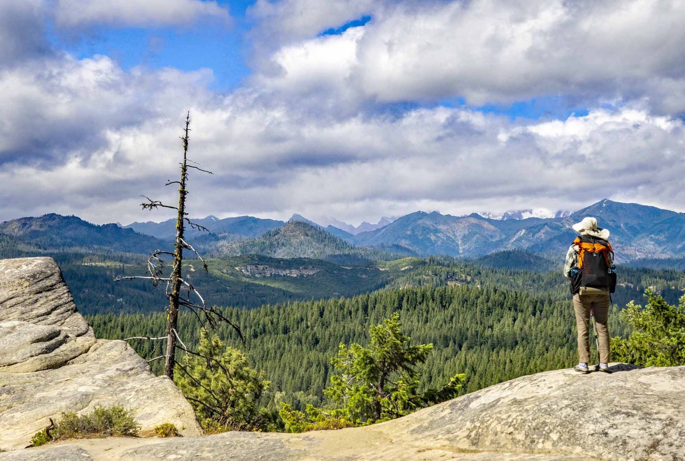 Hiker stands on a rock surface overlooking a valley of evergreen trees and mountain peaks