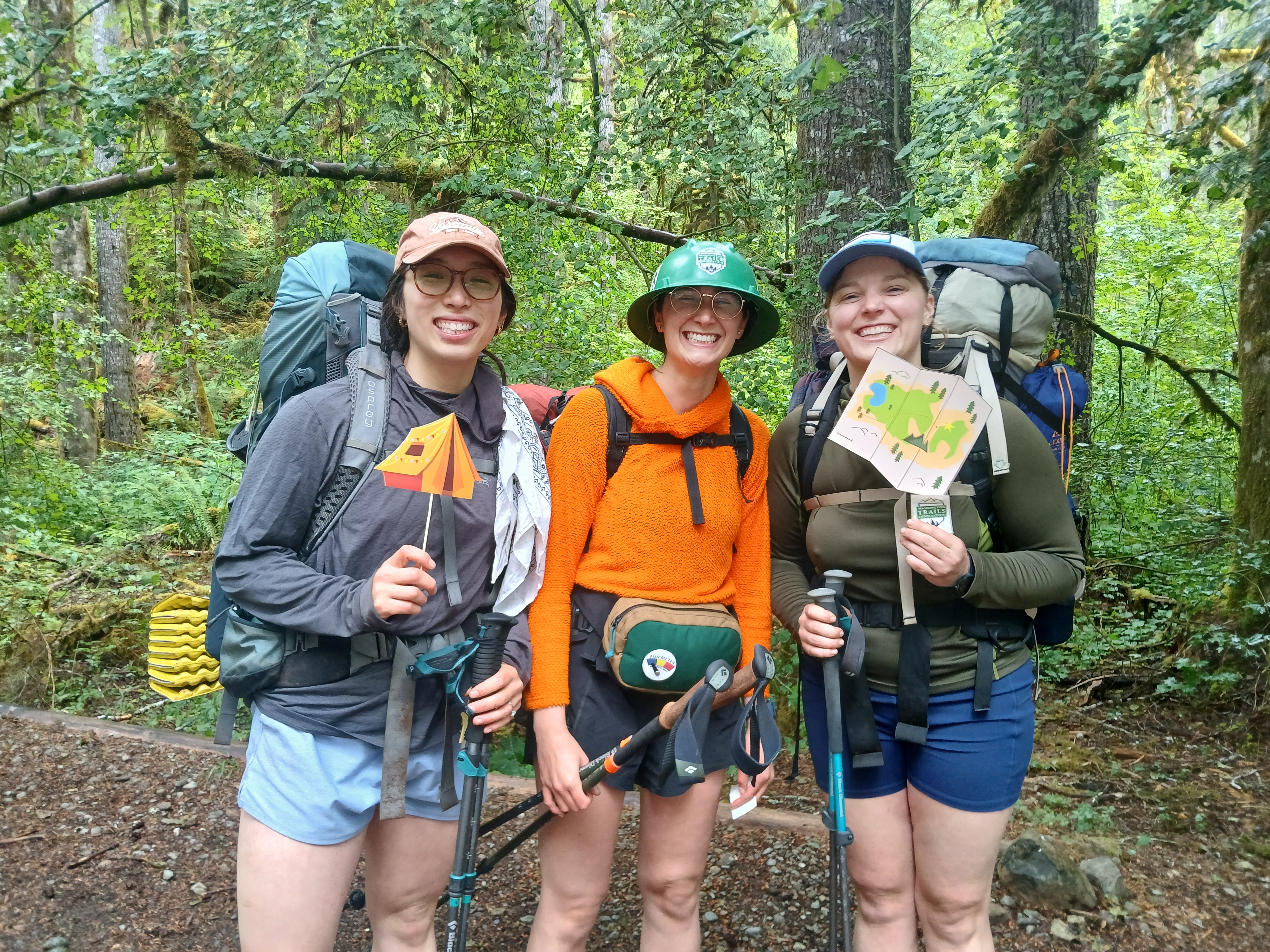 Three smiling hikers in backpacks stand at a trailhead