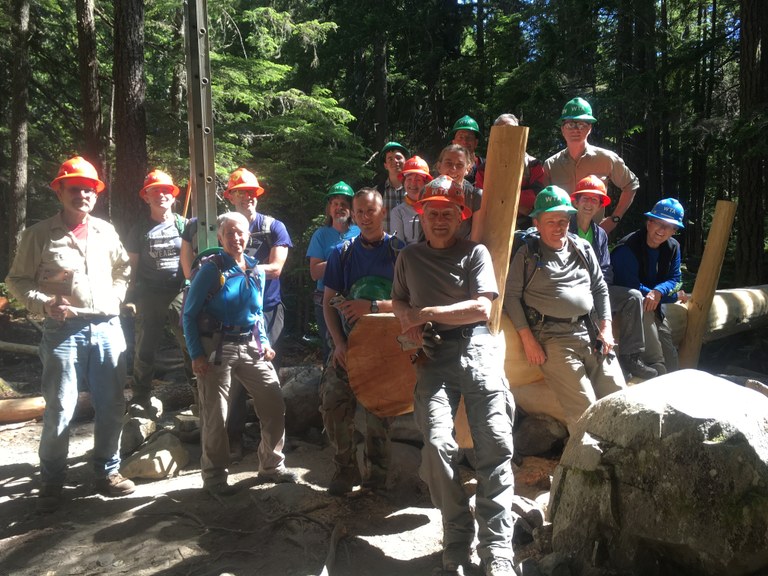 Crew at Dungeness Bridge A group of smiling volunteers sit at one end of a large log bridge in dappled sunlight