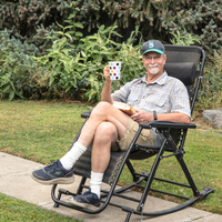 David Hagen Self Potrait Man sits in mesh rocking chair on sidewalk with grass and evergreens in the background. Man is wearing a Seattle Kraken baseball cap, smiling while holding a white coffee mug and book in lap.