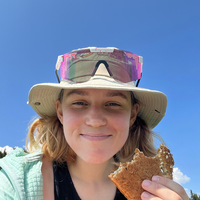 PJ Heusted on trail Women holds piece of bread taking a selfie wearing pink sunglasses and khaki hat on top of head. Blue skies in background.