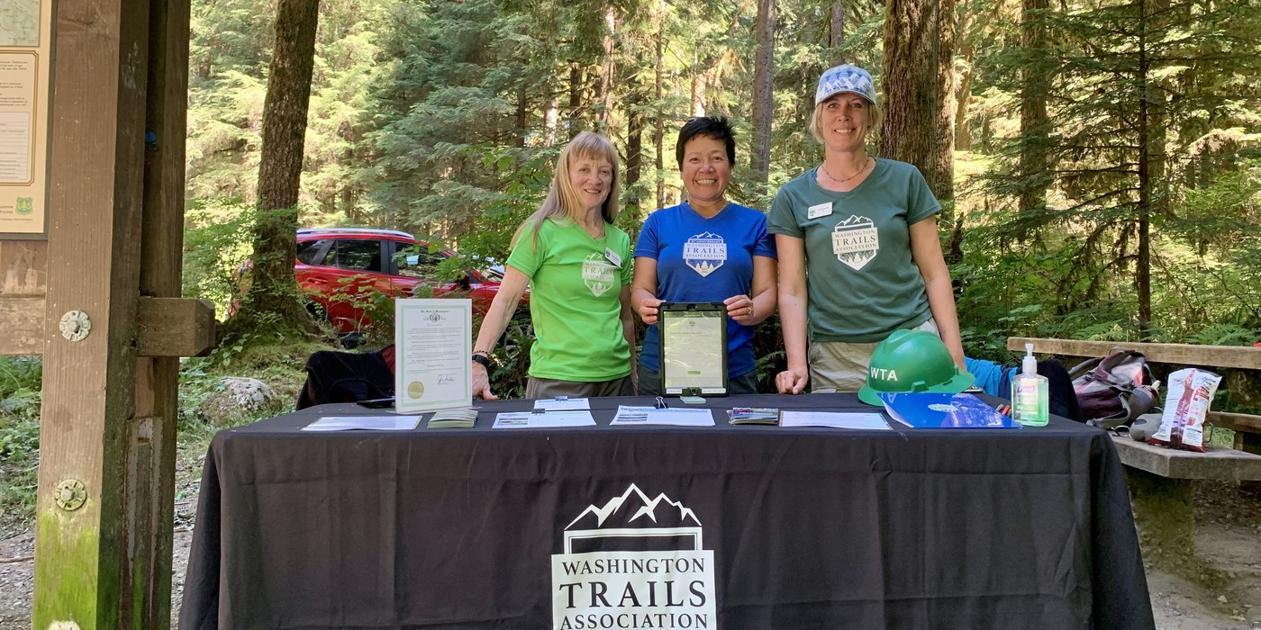 3 volunteer ambassadors stand at an outreach table at a trailhead