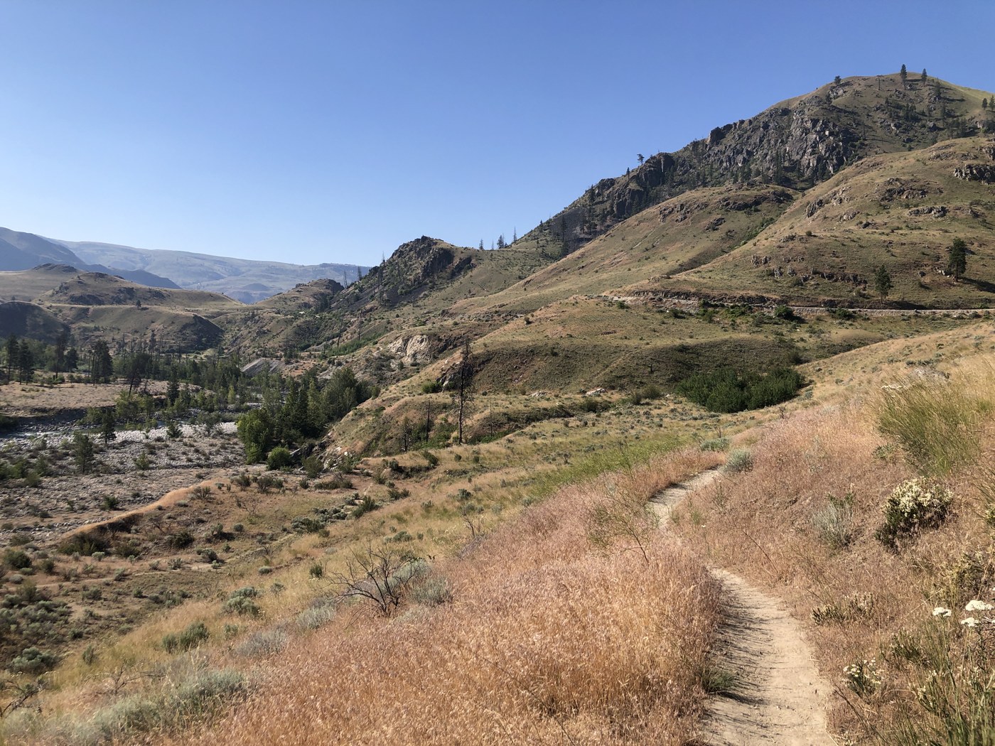 Trail winds out of view on grassy, desert mountains.  Few trees scattered across landscape as river rolls on the far lefthand side of photo.