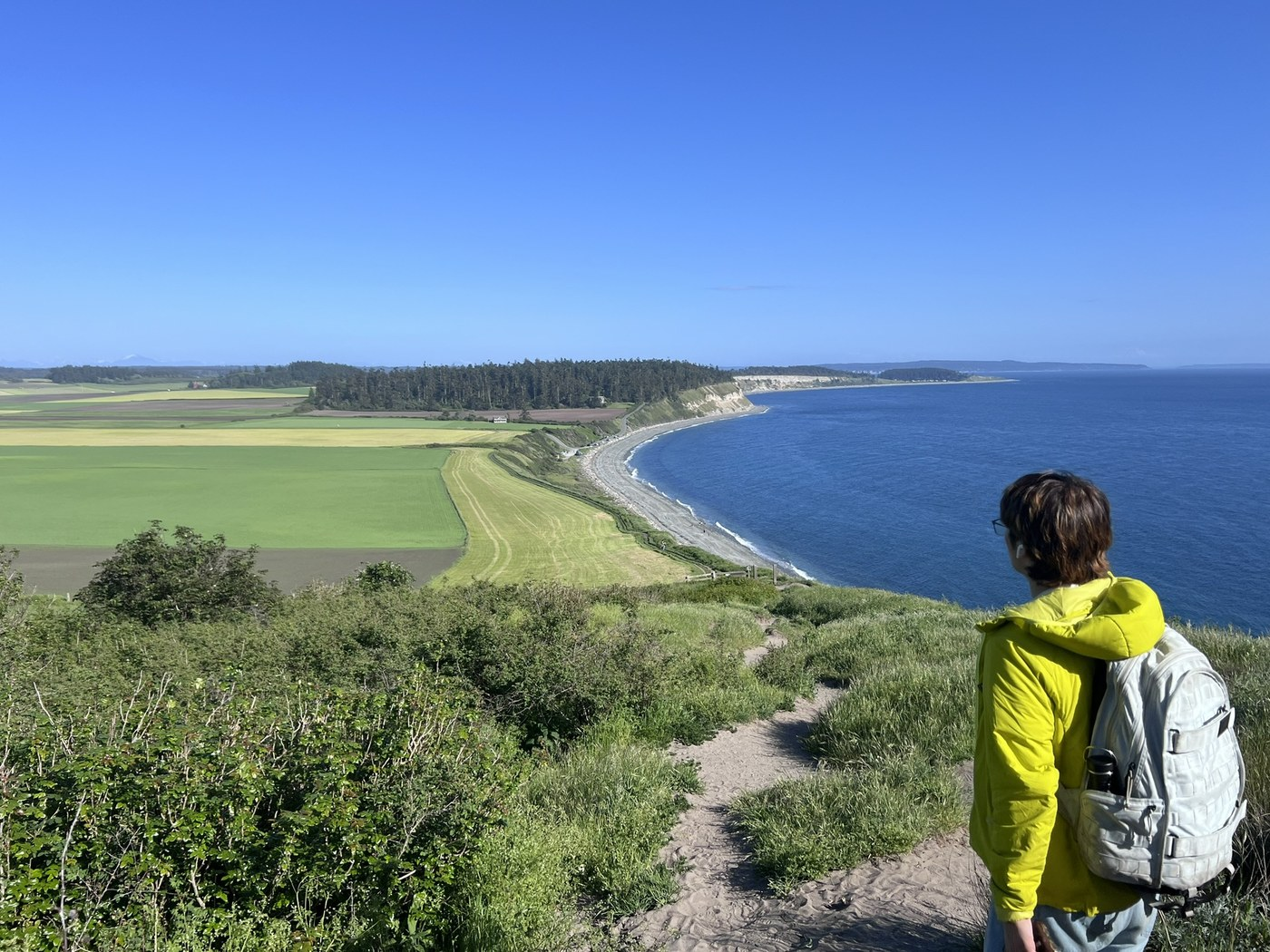 Person in bright green jacket with grey backpack on right facing the water. Sandy trail in between shrubs overlooking ocean and beach to right and green open praries to left. 