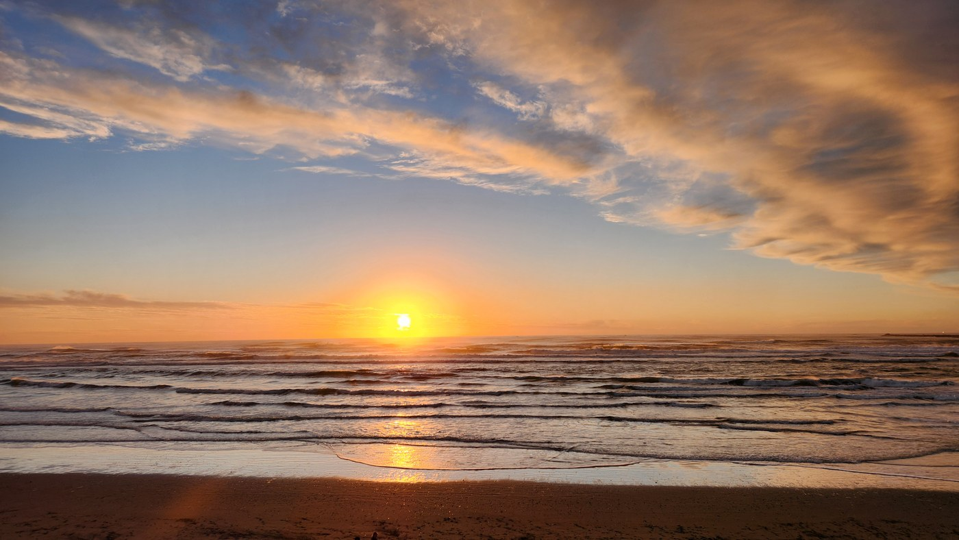 Orange and blue sunset over ocean. Small waves break on shore. Some clouds scattered in the sky.