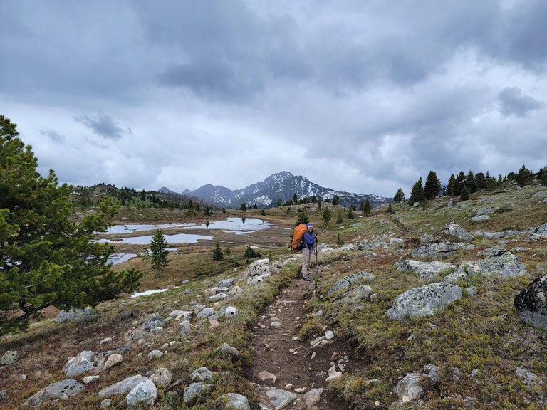 A hiker with a backpacking pack on a trail in the Pasayten Wilderness looking the photographer with lakes and a mountain in the background on a cloudy day. Photo by KellBell.