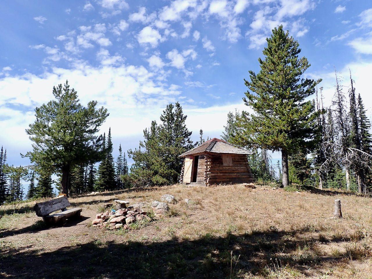 A cute little cabin sits at the top of a sparsely-treed hill agains a backdrop of blue, cloud-scudded sky.