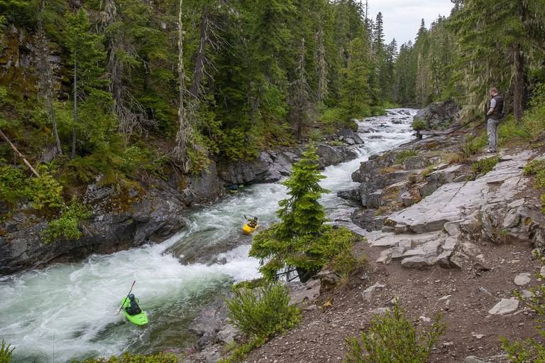 Two kayakers in a green and yellow kayak come down a rushing Class 3 river as a hiker on the banks watches.