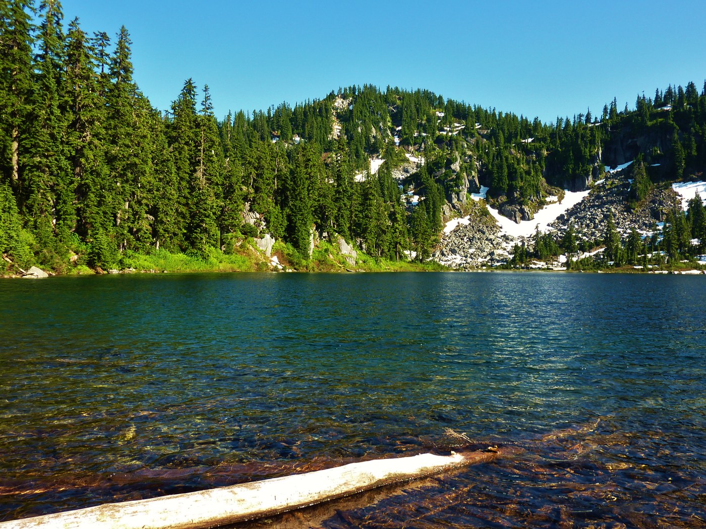 A bright blue lake is framed by green, teed covered hills in the background which are dotted with snow patched.