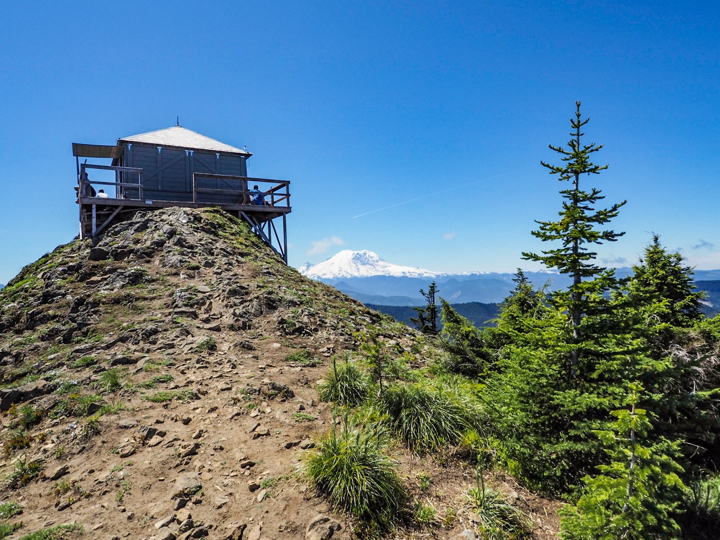 A fire lookout tower is perched on a rocky hill overlooking a forest filled valley.