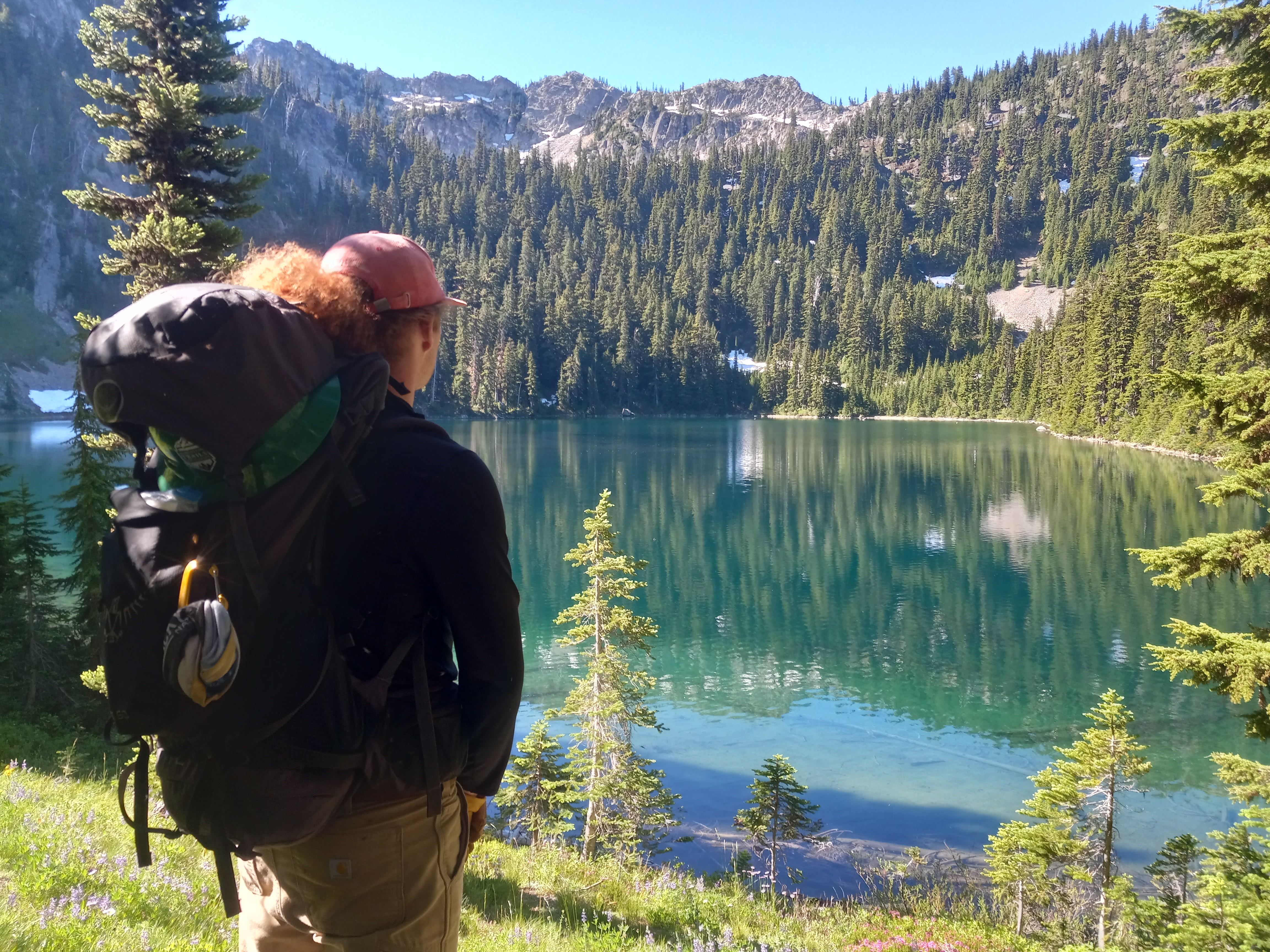 A backpacker stands in the left foreground admiring a large blue-green lake.