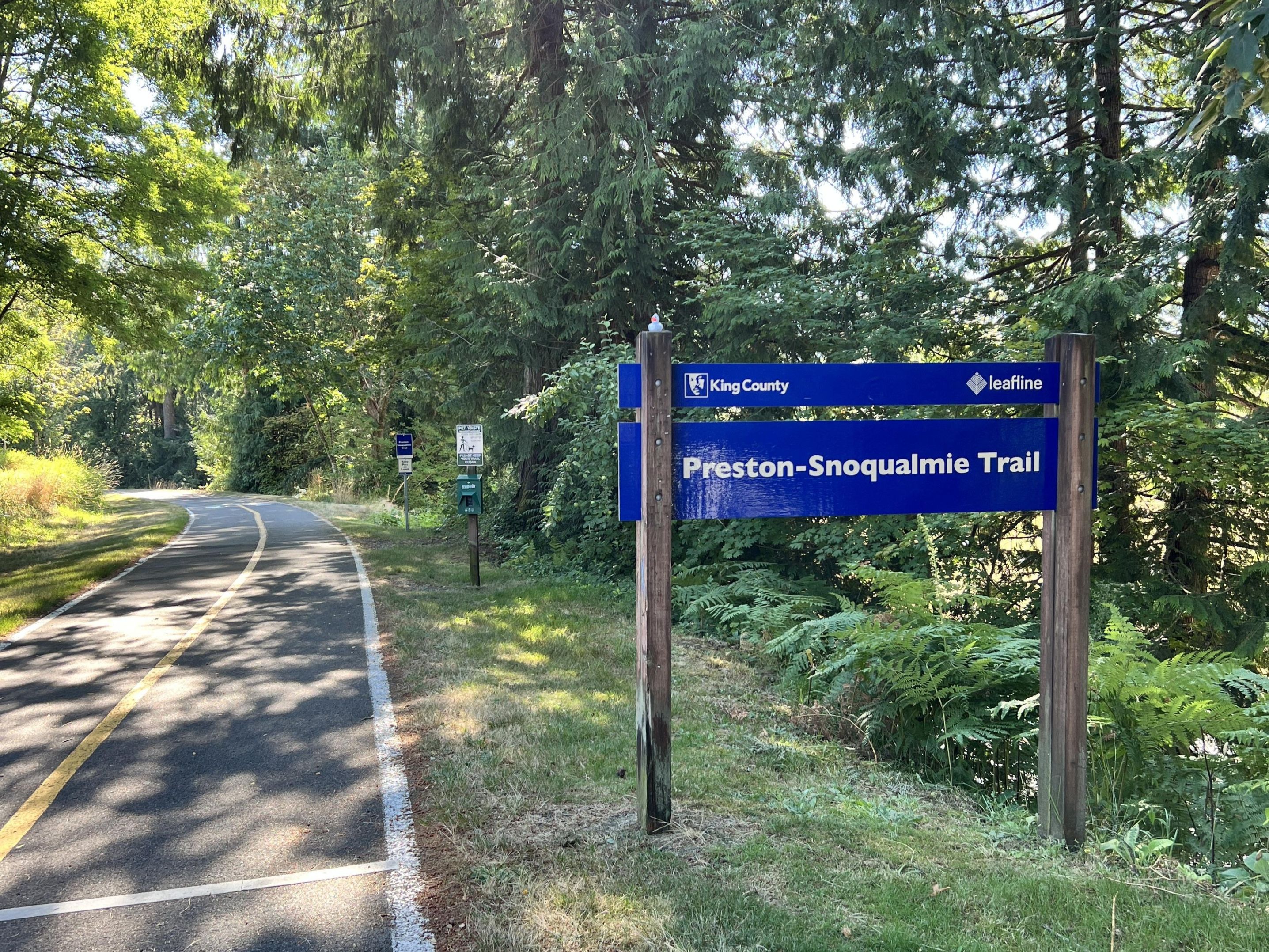 A sign for the Preston-Snoqualmie Trail, with the trail in the background. Photo by BeaverDawg.