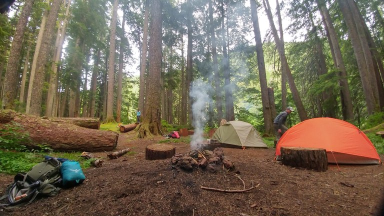 A campsite with tents, a campfire and a couple of people enjoying the area. 