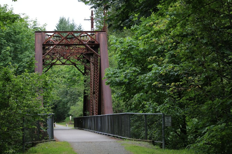 The paved surface of the Cedar River Trail leads to a rusty bridge over the Cedar River. 