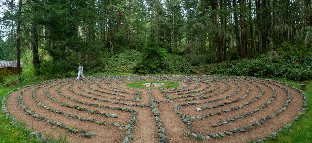A teen walks a rock-lined labyrinth in a green clearing. 