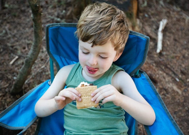 A young boy sits on a camp chair looking at a s'more. Photo by Erynn Allen.