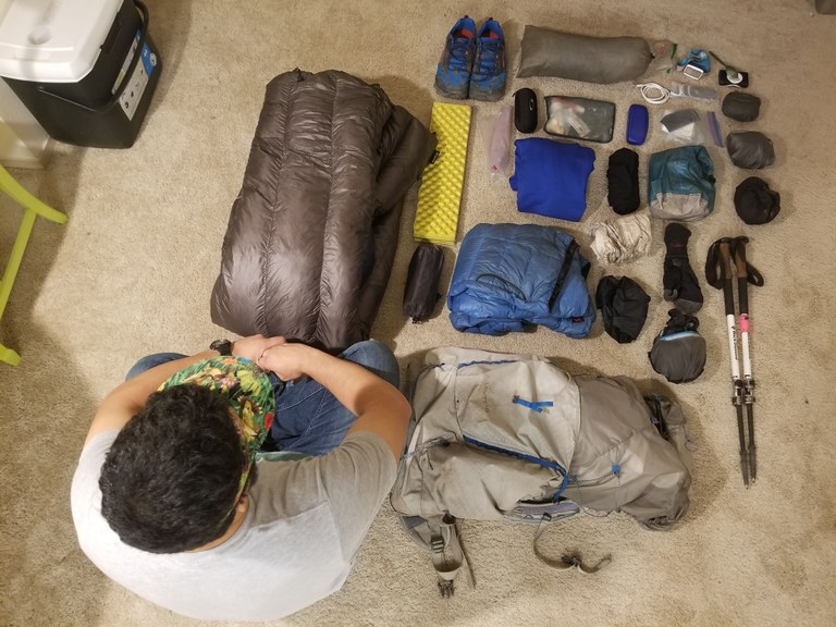 A collection of gear is organized on the floor as a hiker looks it over.