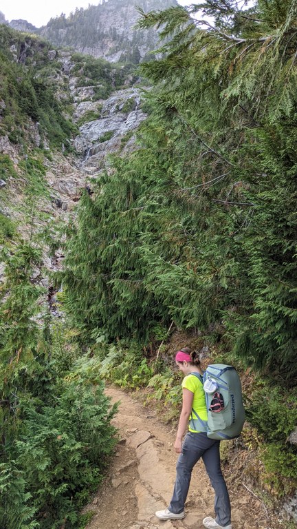 The author strides up a hill on the trail to Gothic Basin while smiling and wearing a green pack. 