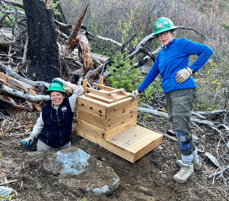 Two WTA backcountry volunteers pose next to a newly made privy box. One of them stands inside the hole the privy will so go over.