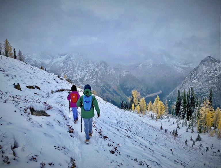 Two hikers walk along a trail covered lightly in snow. 