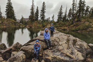 Three hikers lounge on a rock on a cloudy day wearing rain gear.
