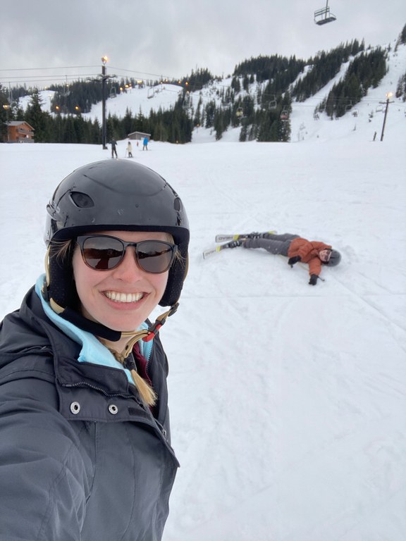 The author's friend takes a break from skiing to take a selfie with the author laying flat in the snow after a fall. Both grin. 