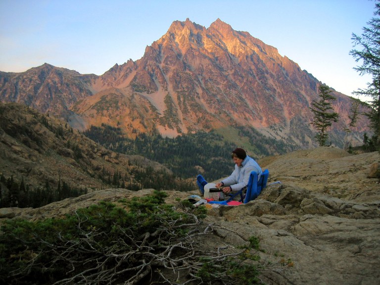 A hiker prepares dinner on a stove in the wilderness. The sun sets on a dramatic mountain face in the background.