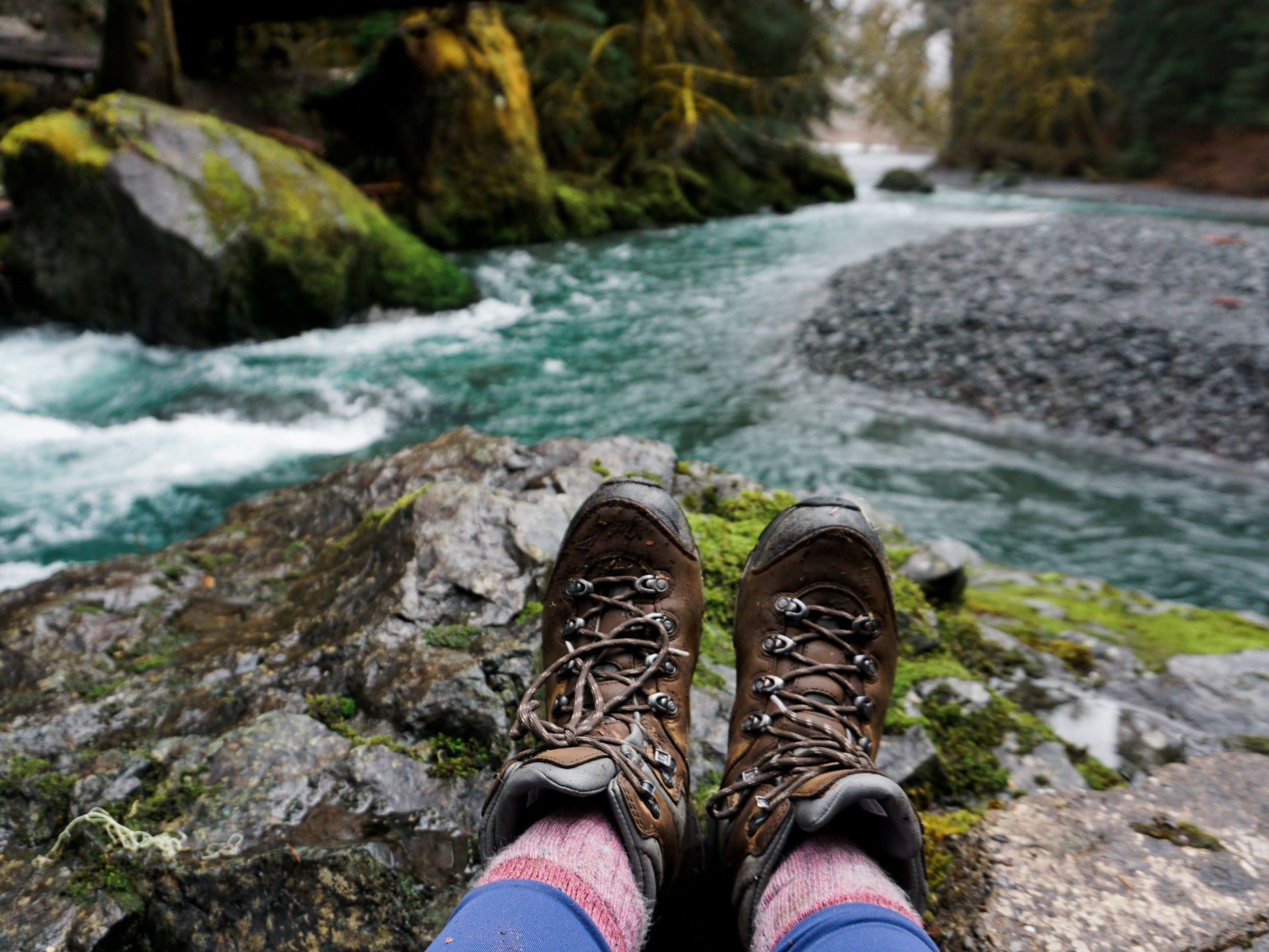 A hiker sits with their feet out in front of them, facing a river.