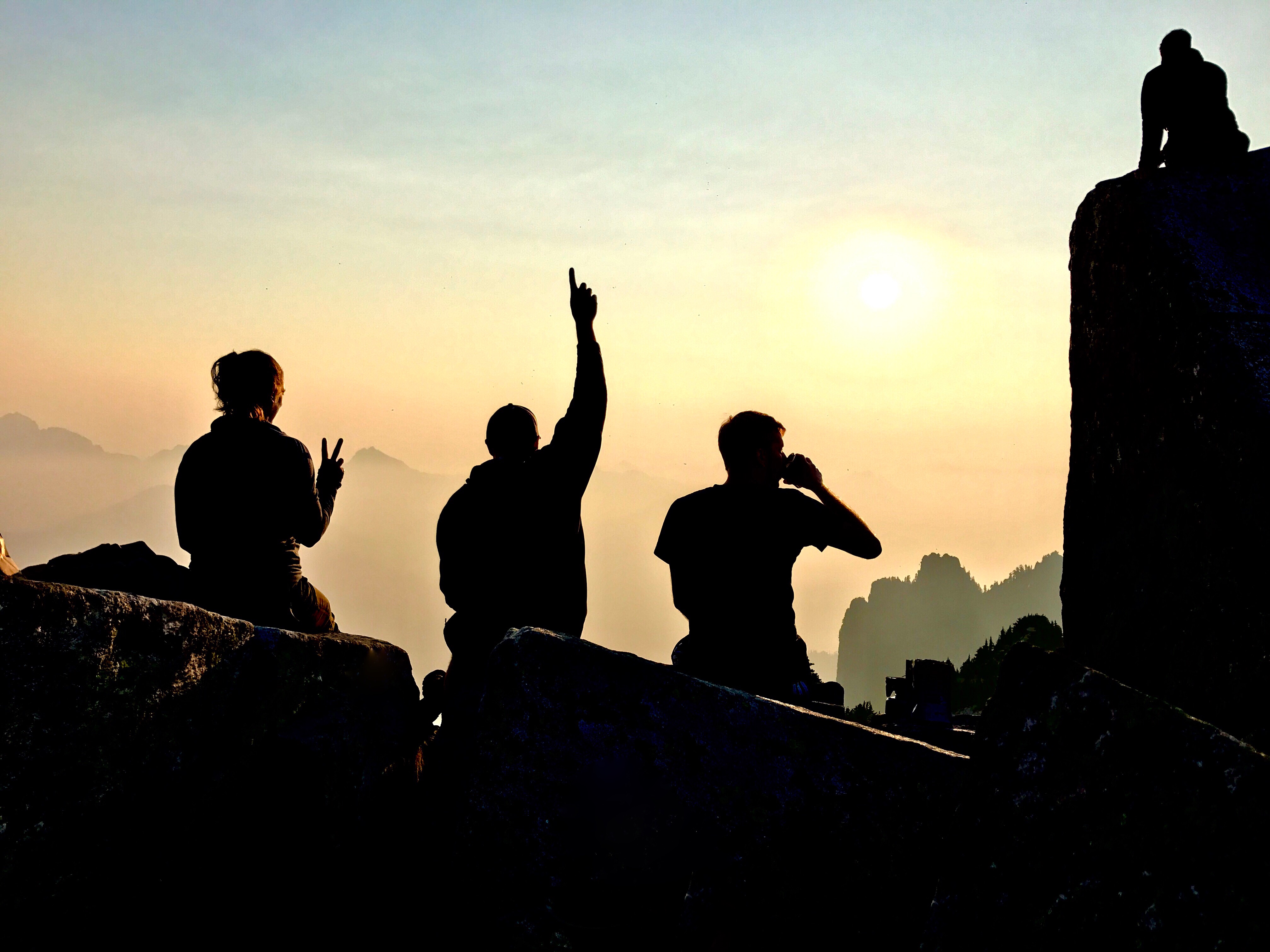 A row of hikers face away from camera and toward the setting sun in the distance. One hiker is making a peace sign with their hand, while another is pointing up at the sky.