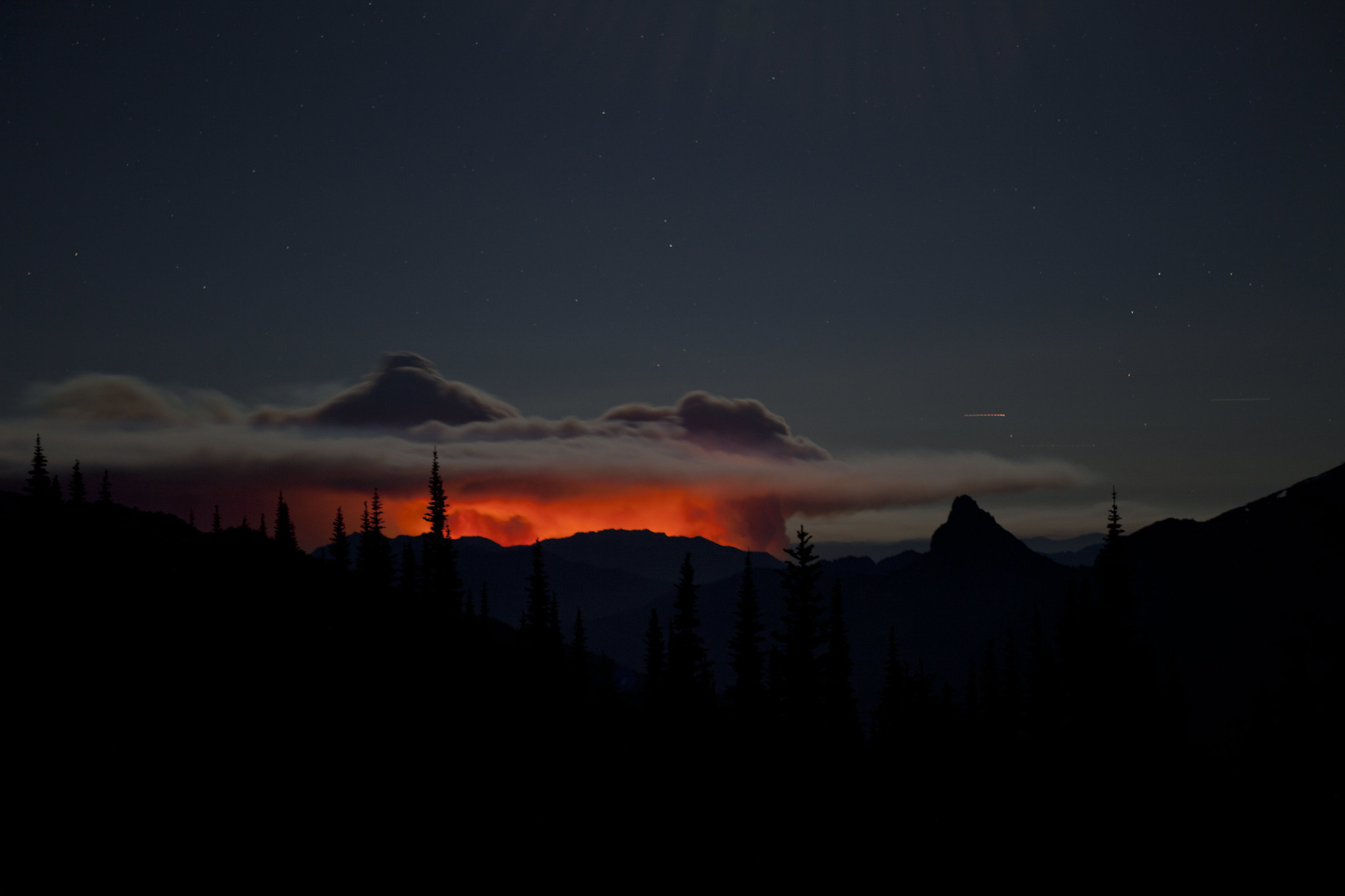 A nighttime view of a glowing red fire burns in the distant forest, with a large smoke plume above it.