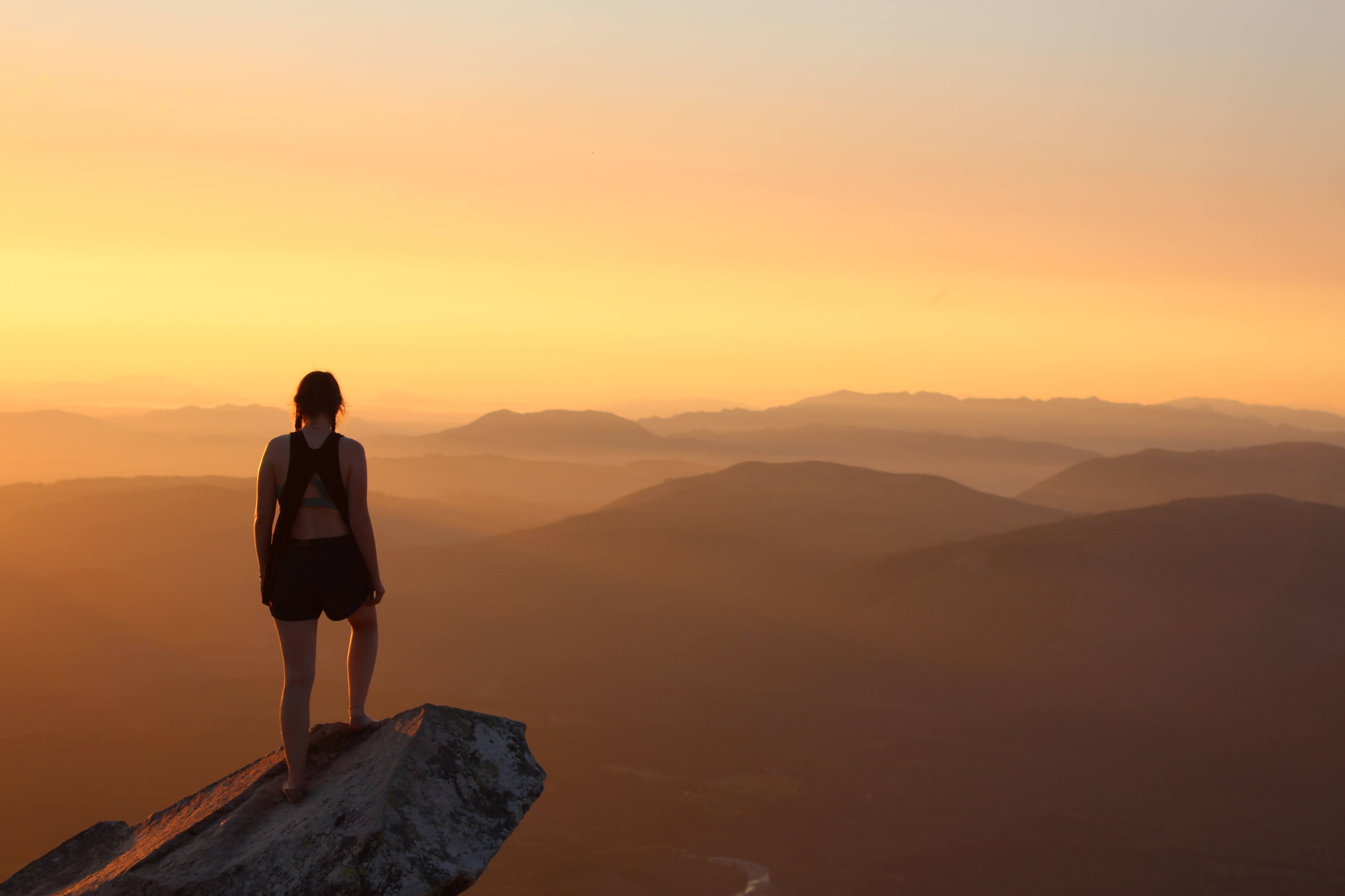 Hikers looks out at skyline, showing layers of mountains, during an orange sunset.