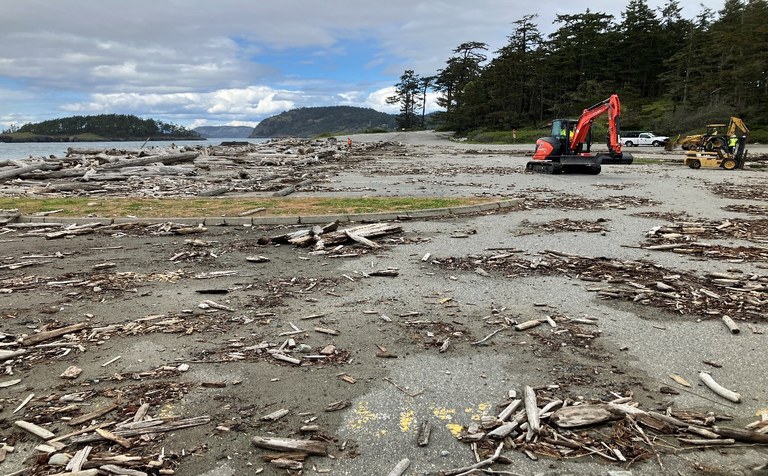 Sand beach littered with debris. Two larger bright construction machines in the background. Blue skies and some water and evergreen trees in the background.