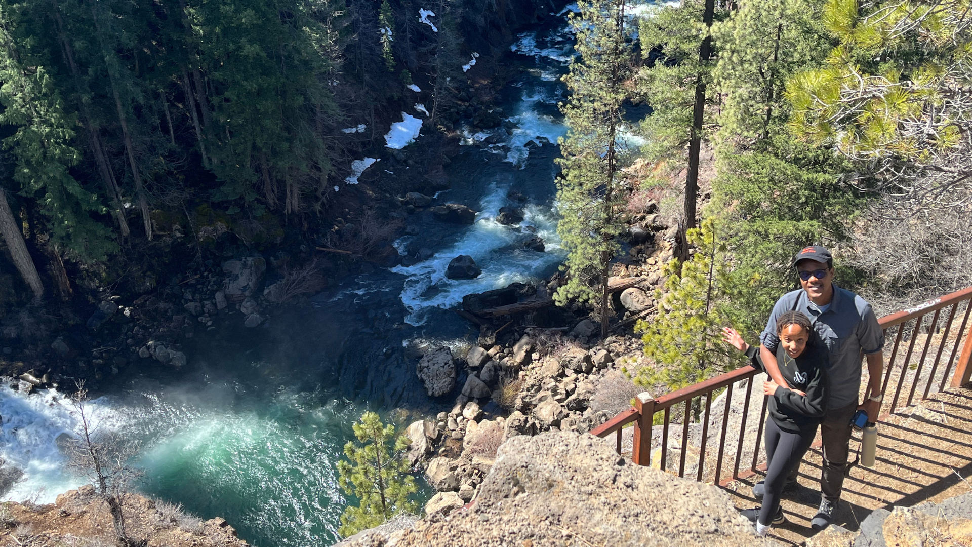 Dr. Ian Munanura with daughter on trail. Photo courtesy of Dr. Ian Munanura. Person stands with child smiling at camera, standing in front of a railing that overlooks a rushing river below.