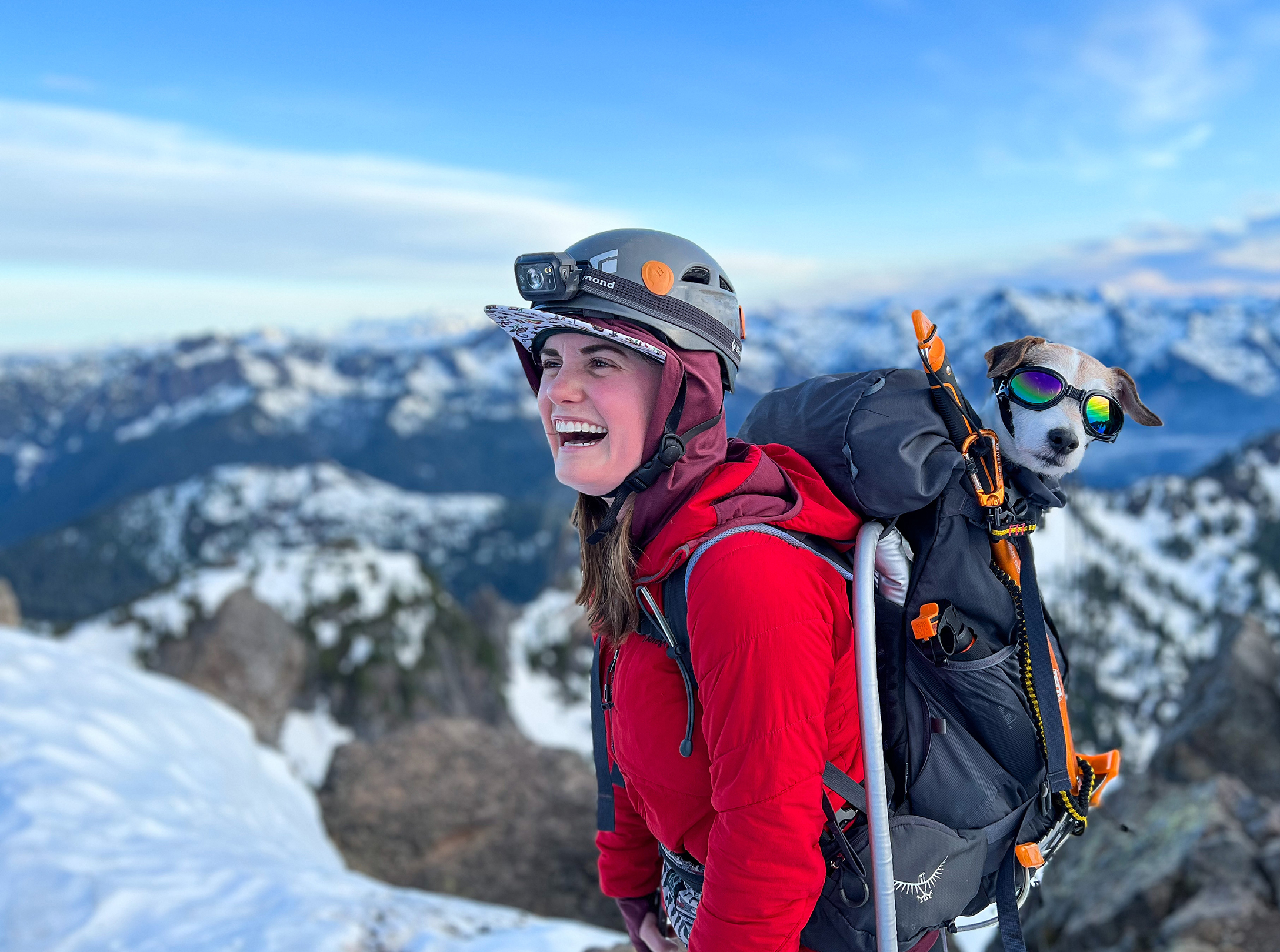 A hiker in a red coat with a dog on her back in a backpack. The dog is wearing doggles. 