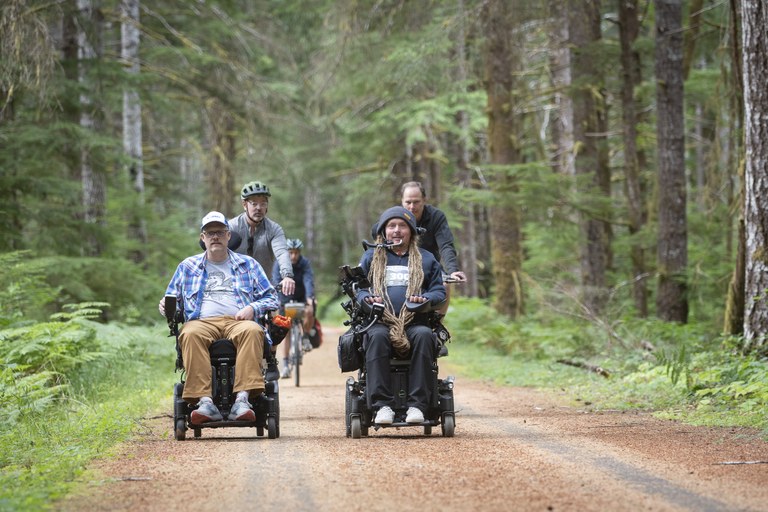 Ian rides on the Olympic Discovery Trail along with Jon Snyder (front left), Gov. Jay Inslee’s outdoor recreation and economic development senior policy advisor, who joined Ian and other participants for 5 miles in a wheelchair during Sea to Sound. Two hikers in wheelchairs ride down a forested trail with three cyclists behind them.