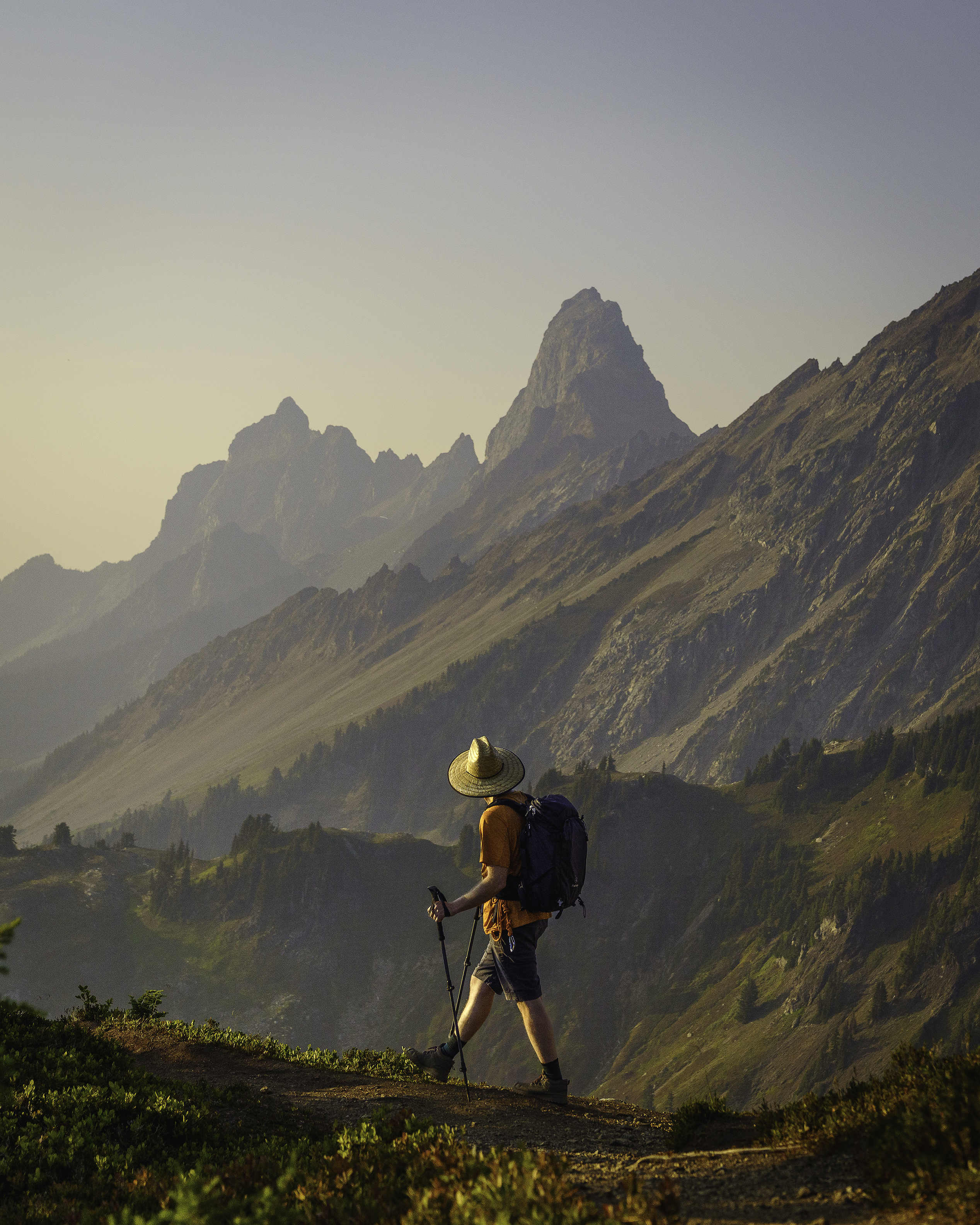 A hiker wearing a broad hat hikes in front of pointy mountain peaks. 