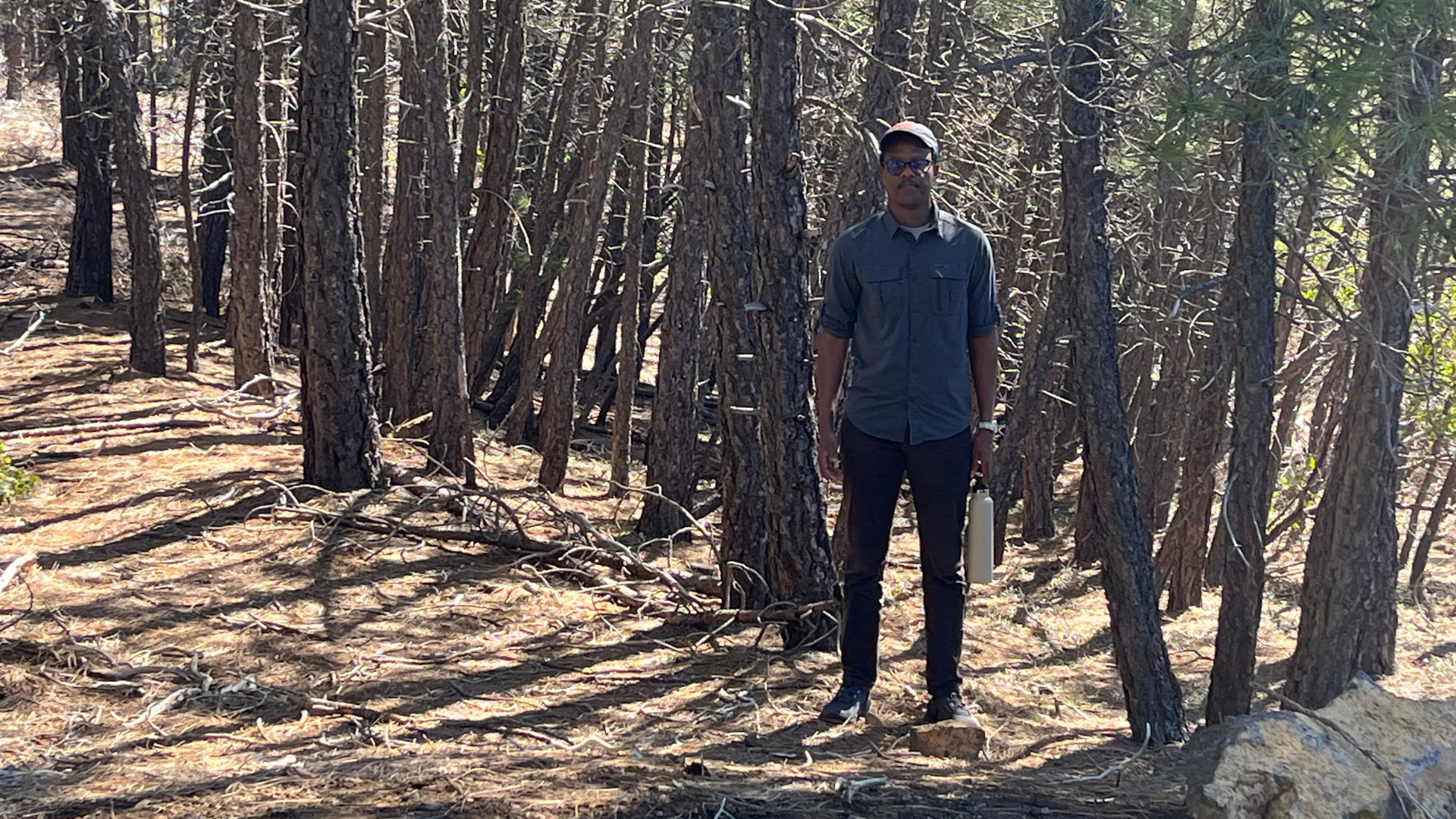 Man wearing baseball cap and blue button down stands amoungst trees on trail holding a water bottle.
