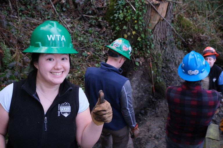One person with green helmet and thumbs up smiles at camera as two other people work in the background.