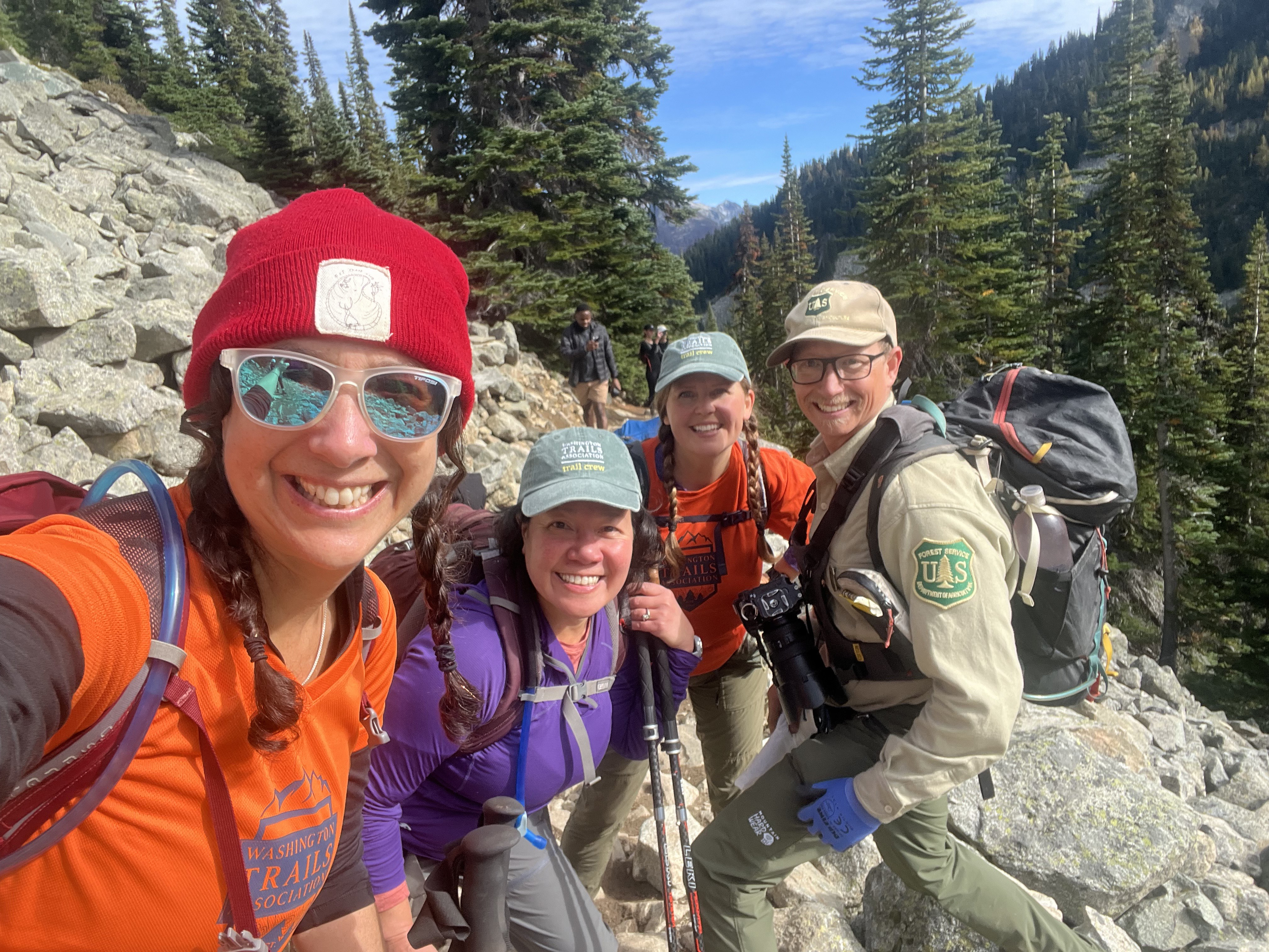 Ambassadors by Nomi Fuchs Photo of four people smiling on rocky trail with blue skies, evergreens, and mountain slopes in the background.
