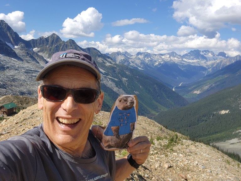 Craig smiles from a mountain vista. He holds up a cut-out of Miles the Marmot, WTA's Hike-a-Thon mascot.