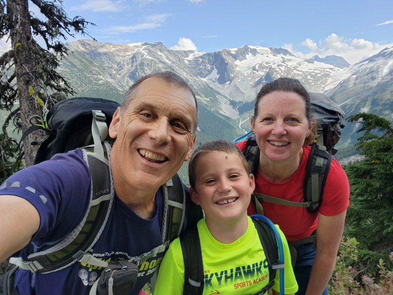 Craig and his family, wearing backpacks, smile in front of a mountain backdrop.