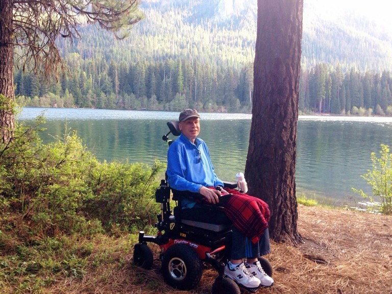 A hiker using a wheelchair in front of a large lake, smiling at the camera.