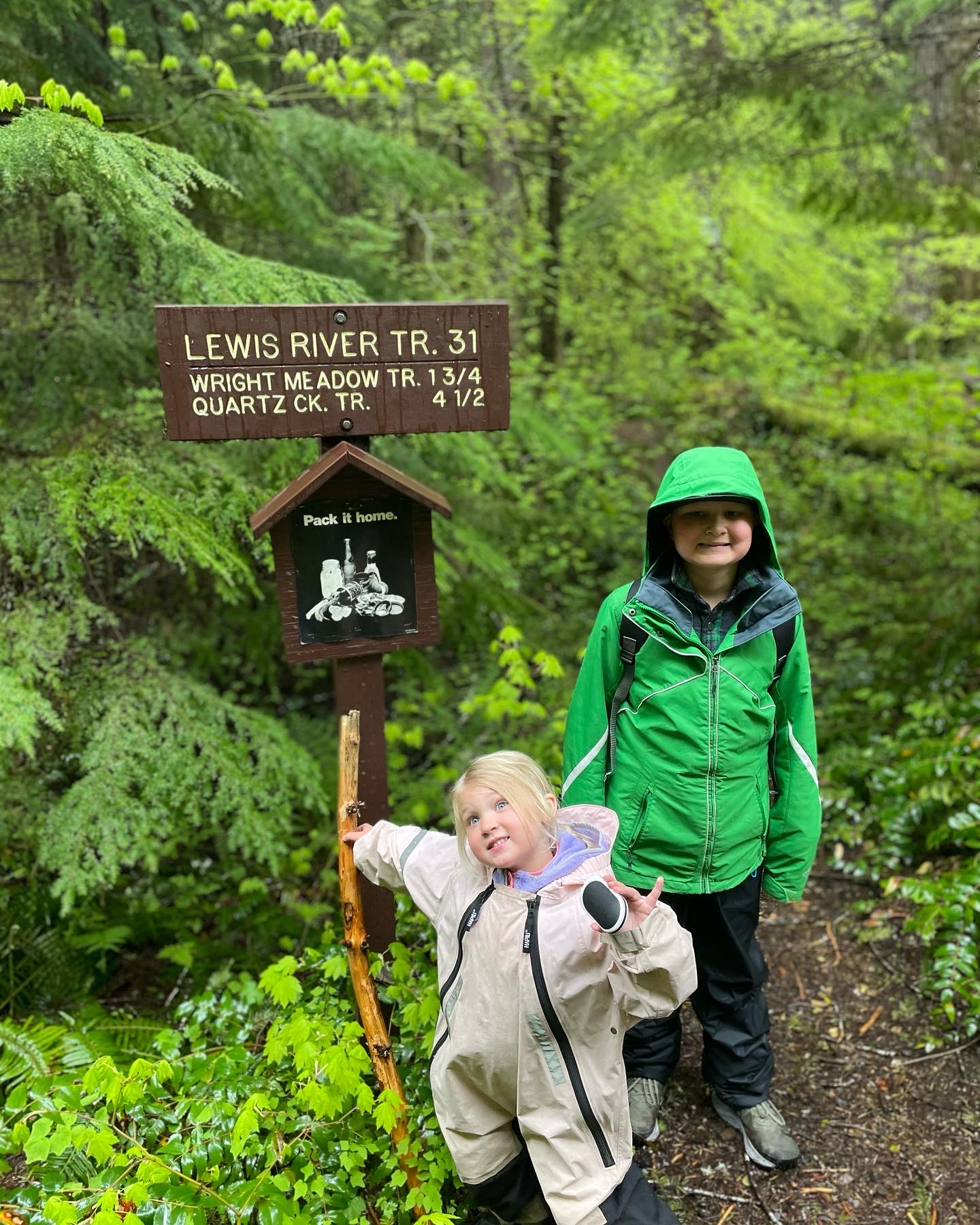 Two kids stand next to a sign for Lewis River, while wearing rain gear. 