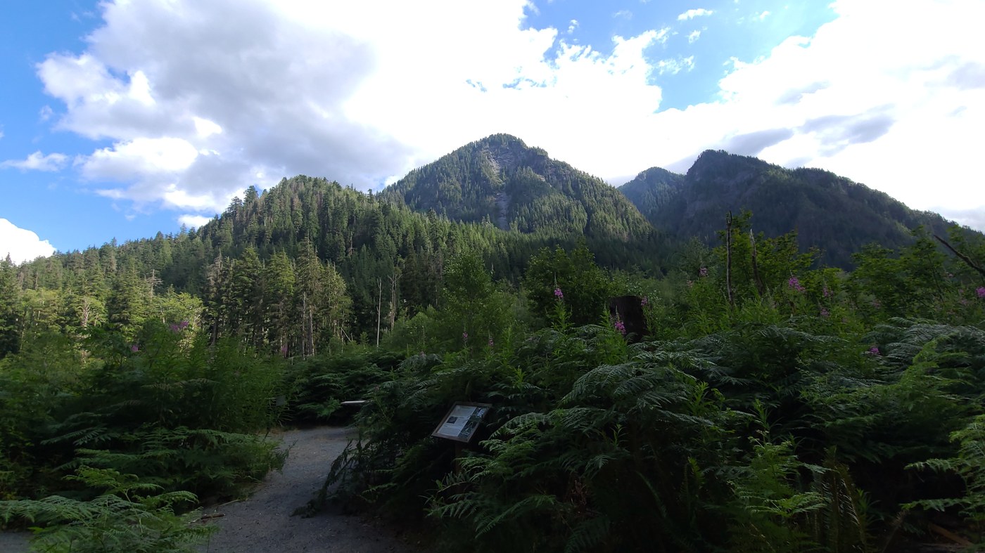 Camp Brown by trip reporter RickyLM Partily cloud skies float above forested mountain peaks. Trail signs and gravel trail at bottom of photo.