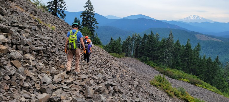 Trail maintainers walk along a trail of talus. In the distance is a volcano.
