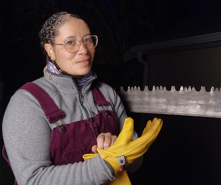 From cutting her first log cookie to helping to restore a popular climbing crag, Crystal’s crosscut saw journey has had a powerful sense of community. Photo by Crystal Hudelson Crystal puts on work gloves while posing in front of her crosscut saw.