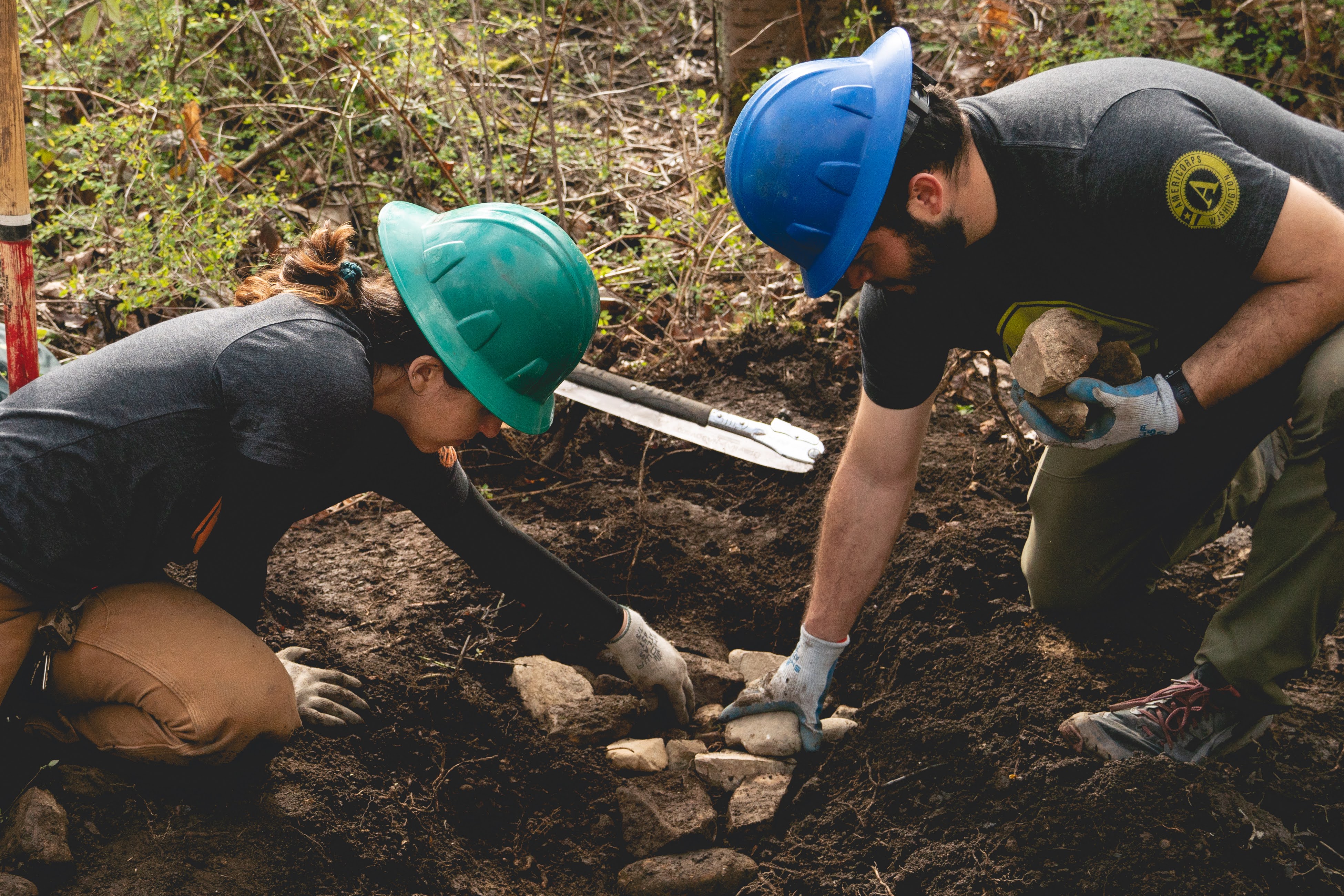 One person in blue hard hat and one in green piecing together rocks on trails.
