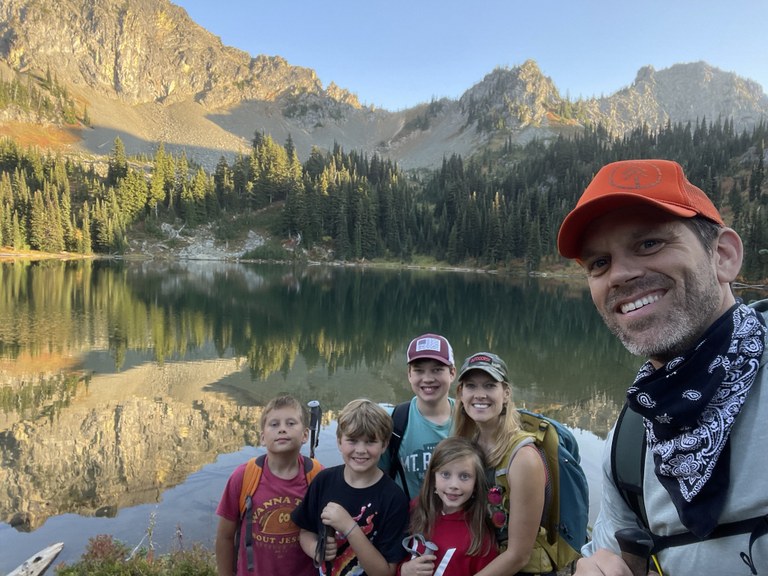 A family of hikers smile next to an alpine lake at golden hour.