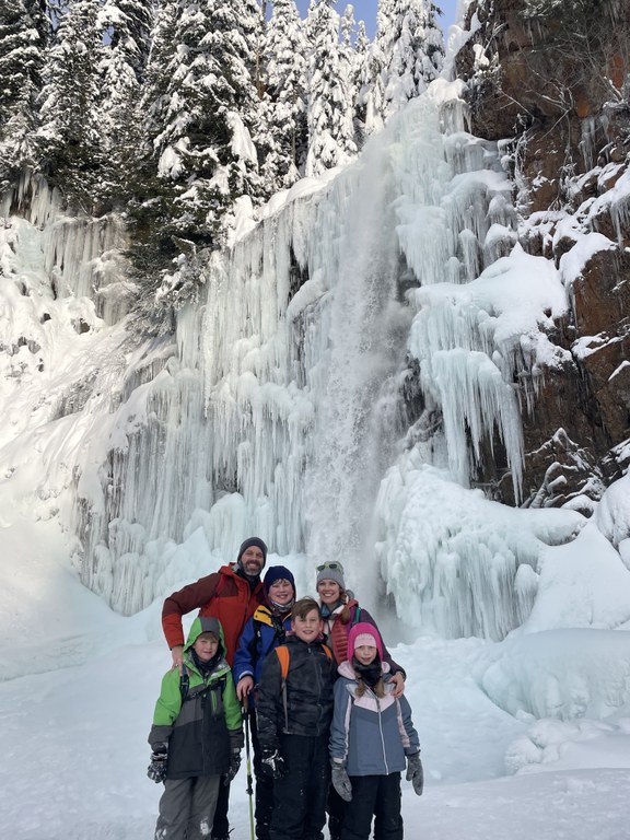 A family in winter apparel smiles in front of a frozen waterfall.